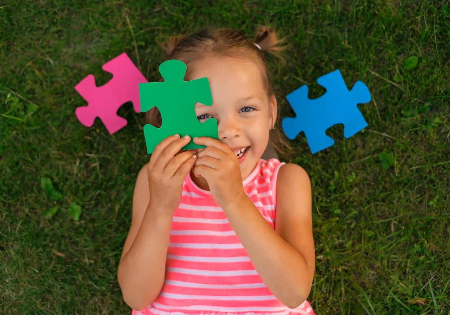 Een schattig mooi kleutermeisje ligt op het gras op het gazon en bedekt haar gezicht met puzzelstukjes terwijl ze vrolijk lacht. Voorbereiding op school, ontwikkeling en onderwijs