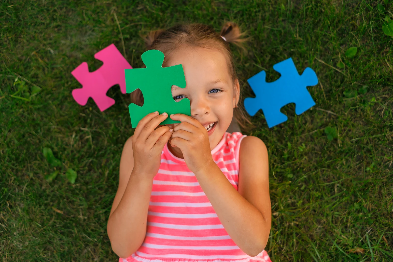 Een schattig mooi kleutermeisje ligt op het gras op het gazon en bedekt haar gezicht met puzzelstukjes terwijl ze vrolijk lacht. Voorbereiding op school, ontwikkeling en onderwijs