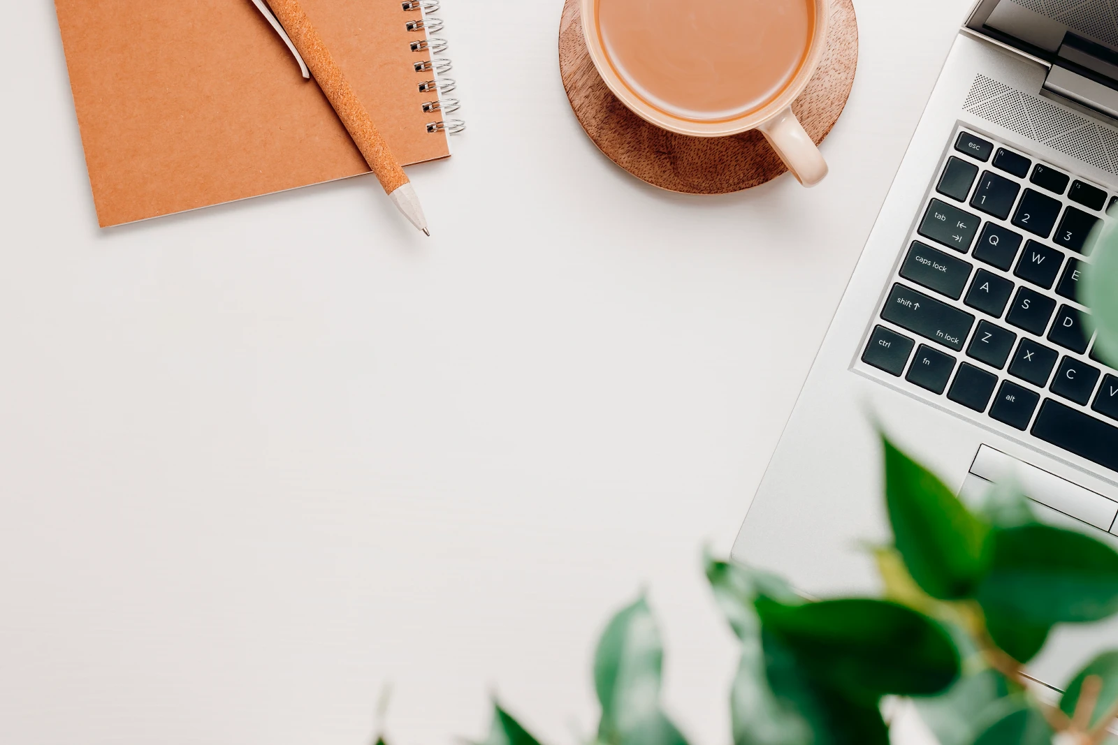 Bureau met kopieerruimte, bovenaanzicht. Laptop, koffie en kantoorbenodigdheden met groene plantenbladeren. Gezellig kantoor aan huis, werk, freelance, creatieve werkplek.